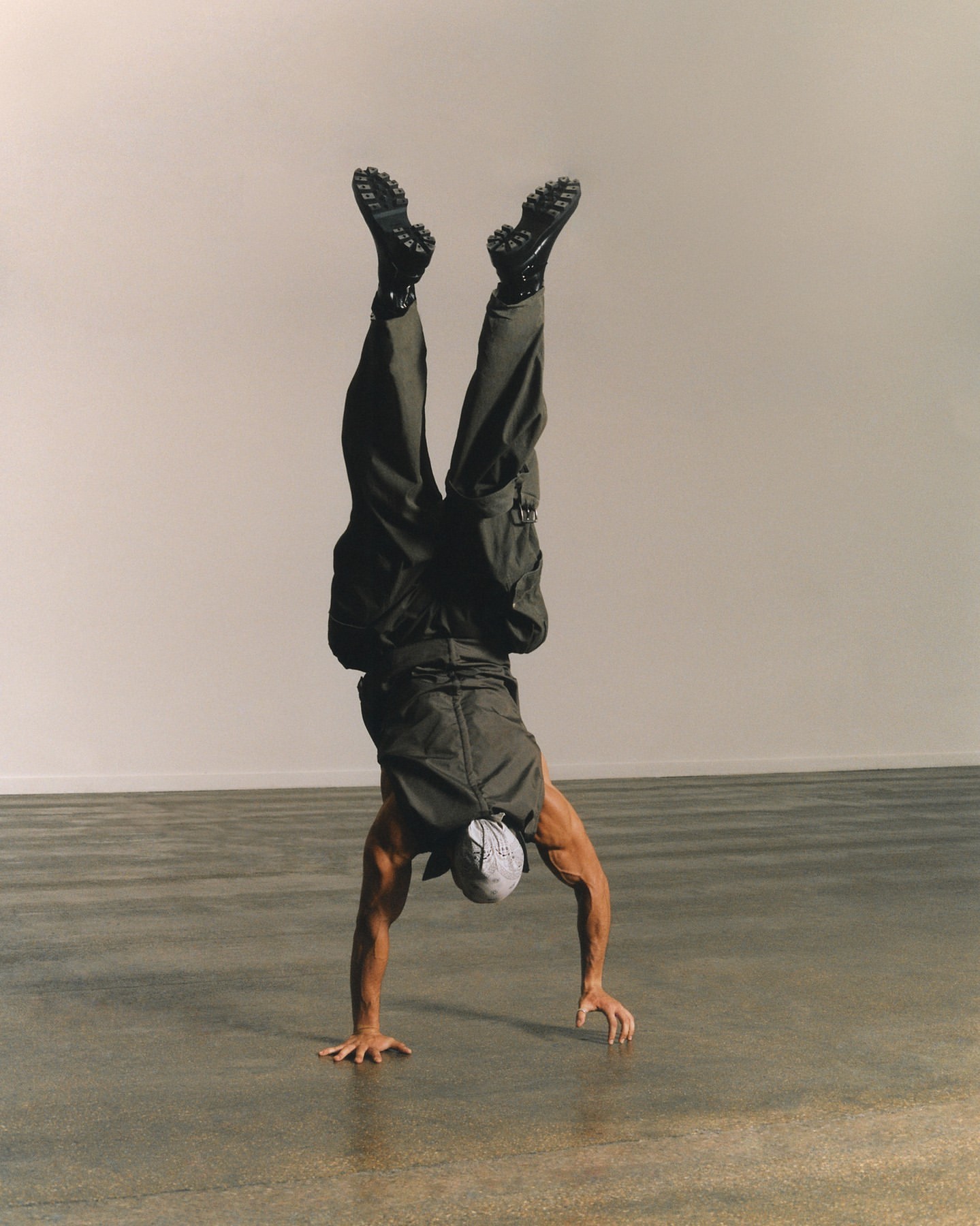Athletic adult does a handstand wearing cargo pants, black boots, and a white head covering in a photo studio setting with neutral light.