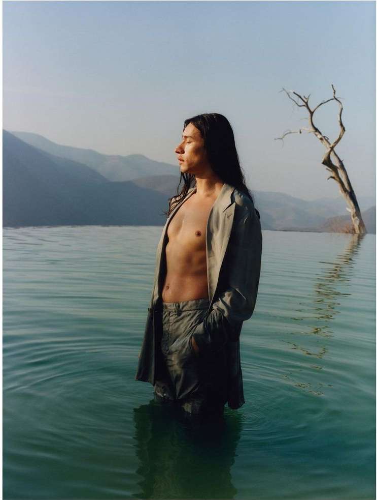 A man stands in water in a light-colored suit with mountains and a tree in the background, illuminated by sunlight during the daytime.