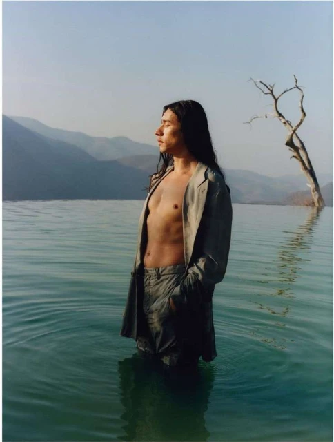 A man stands in water in a light-colored suit with mountains and a tree in the background, illuminated by sunlight during the daytime.