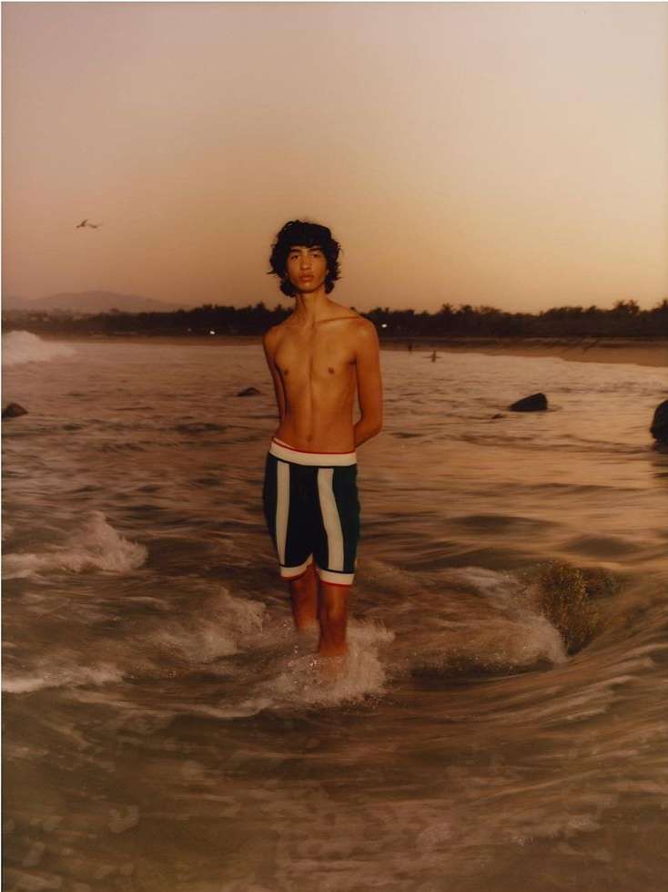 Young man stands in ocean water at sunset wearing striped shorts. The scene captures the serene beach atmosphere and golden light.