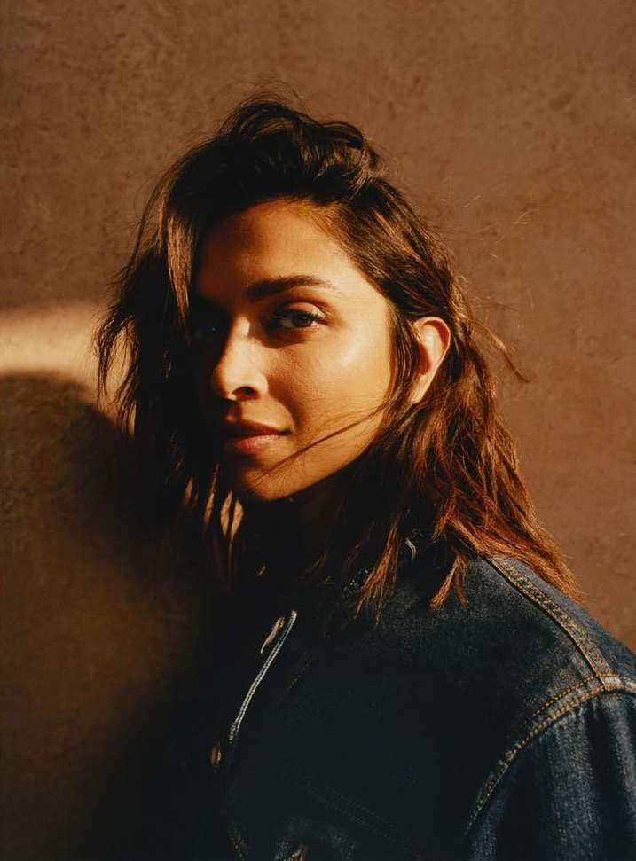 Portrait of an adult woman with brown hair wearing a dark denim jacket in front of a textured brown backdrop with soft lighting.