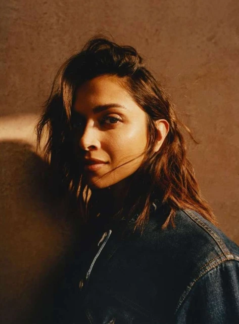 Portrait of an adult woman with brown hair wearing a dark denim jacket in front of a textured brown backdrop with soft lighting.