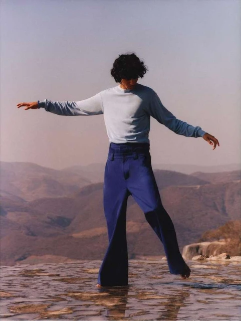 An adult stands on a travertine formation in blue jeans and a long-sleeve shirt. The background includes mountains under a clear sky.