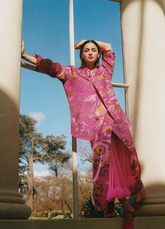 Woman poses in a pink floral pantsuit with white window frame and outdoor background featuring trees and blue sky on a sunny day.