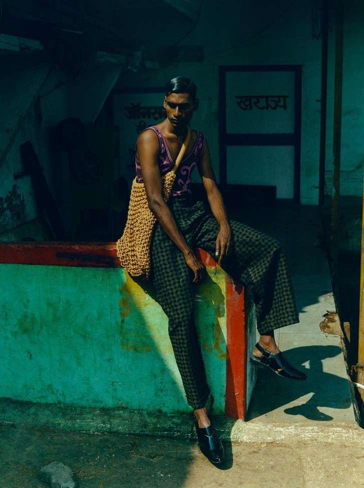 A dark-skinned adult male in a patterned tank top and pants sits on a ledge with a knitted bag slung over his shoulder in urban setting.