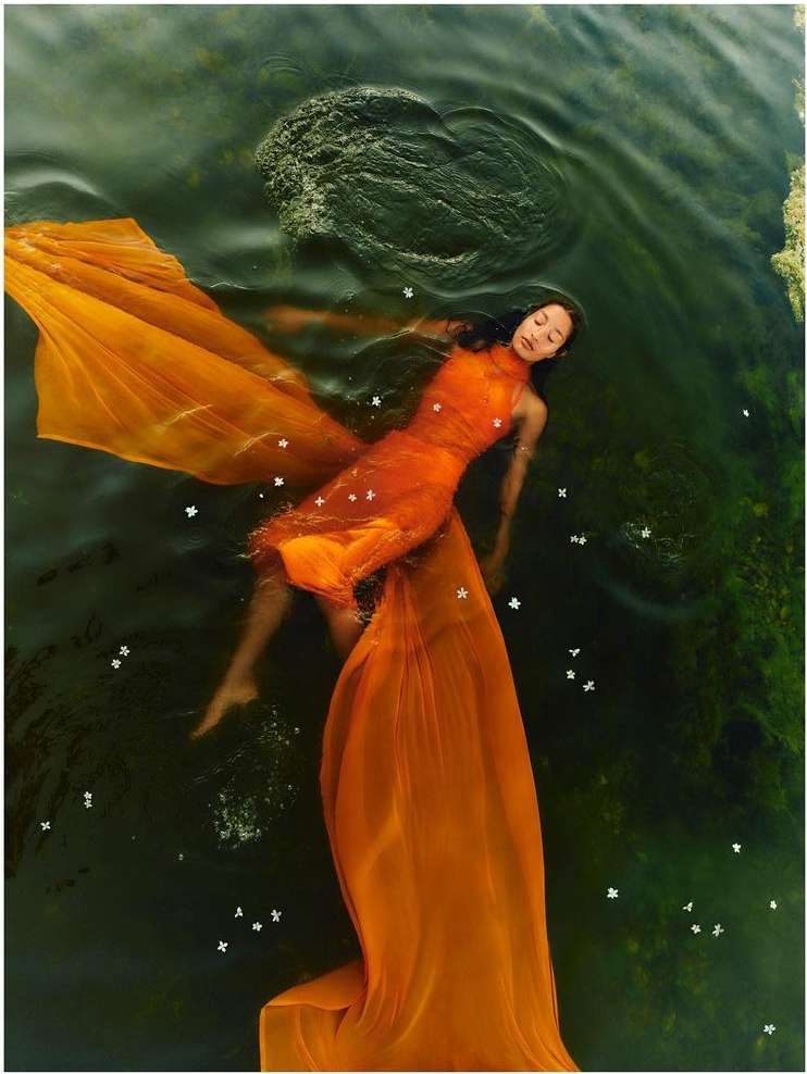 Woman floats in dark water wearing orange dress. Dreamy underwater fashion portrait with flowing fabric and tranquil expression.