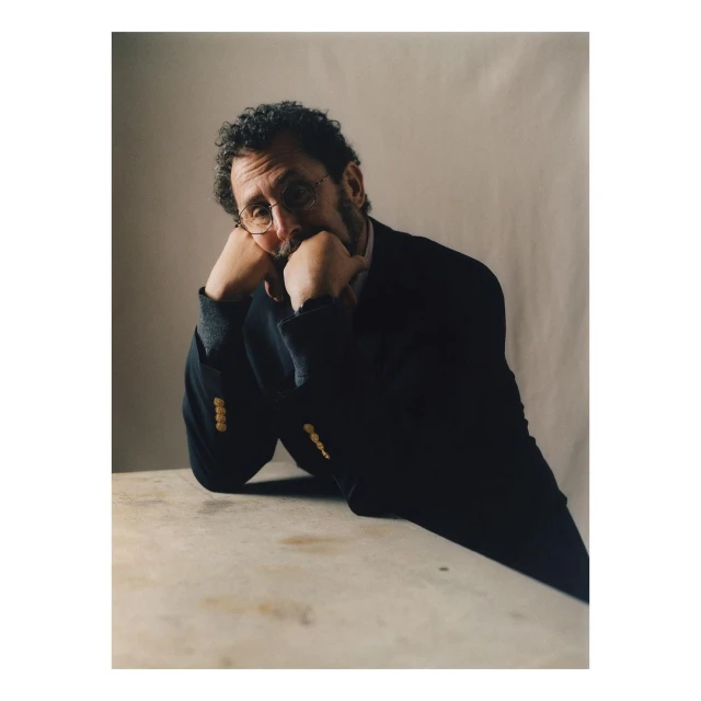 Portrait of an adult man in a blazer with his hands resting on a table, featuring indoor studio lighting and a soft, neutral backdrop.