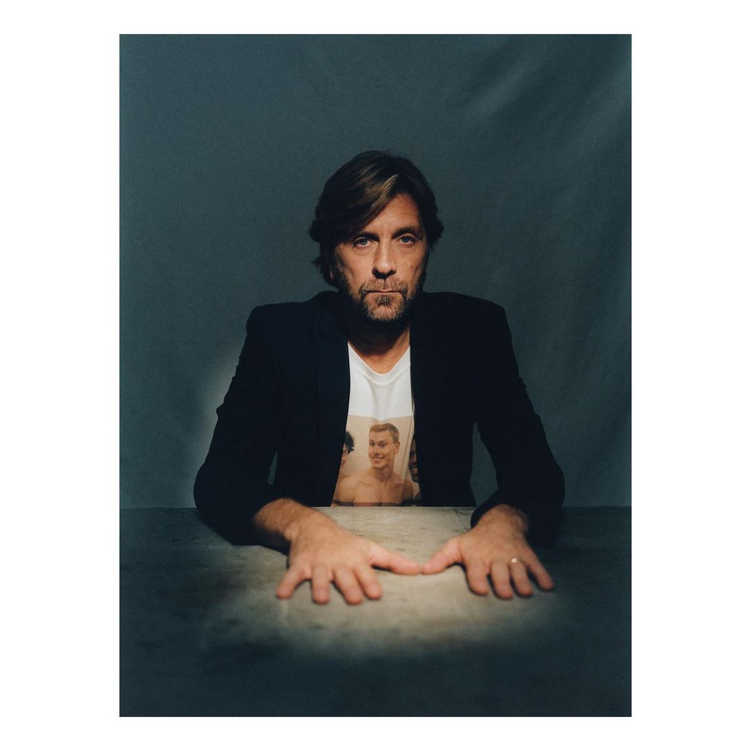 Portrait of a man with dark hair wearing a black jacket over a graphic t-shirt, resting hands on a table.