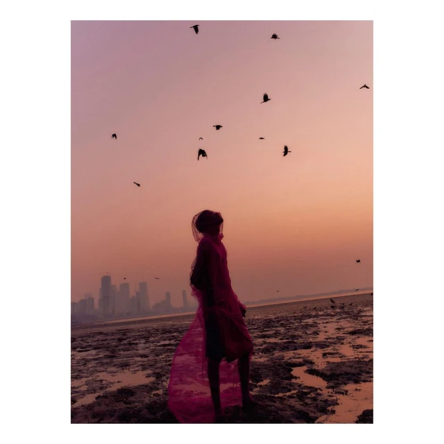 Woman draped in red fabric stands on muddy beach at sunset. Birds fly overhead, distant city skyline. Moody, atmospheric shot.