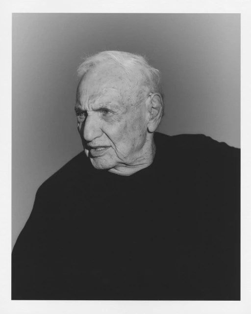 Monochromatic portrait of an elderly man with white hair in a black sweater against a light gray background, looking off-camera.