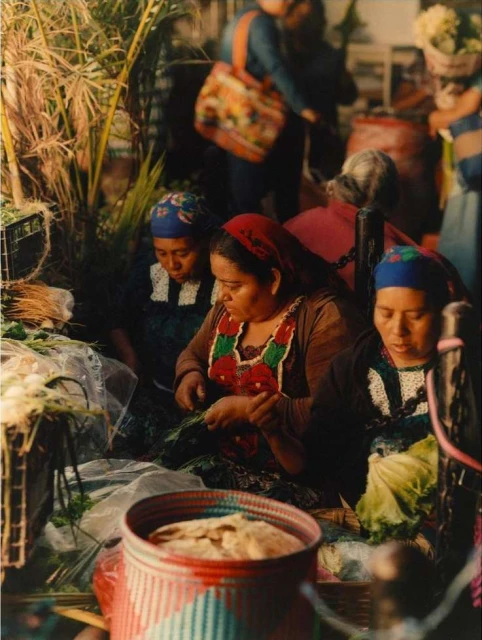 Three adult females in traditional clothing at an outdoor market, with food in a basket and greenery in the background, captured with natural light.