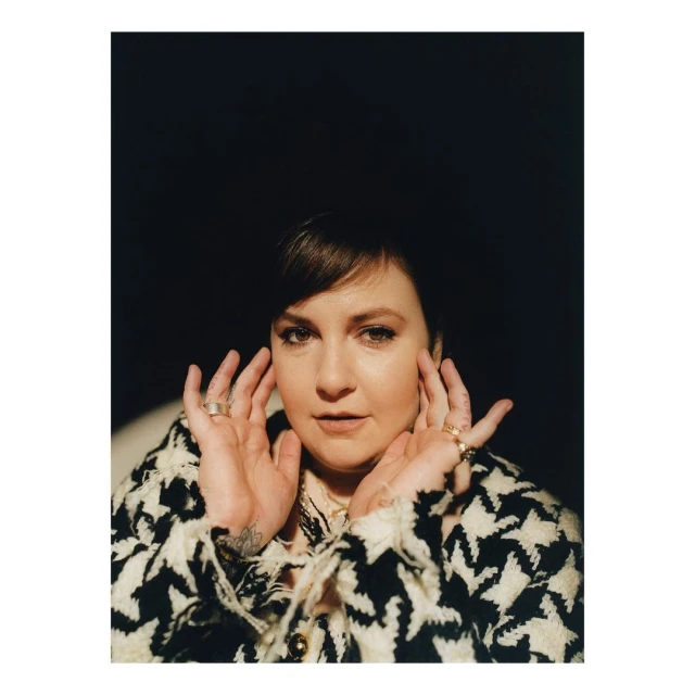 Portrait of an adult woman with hands near her face, wearing a black and white houndstooth coat in a dimly lit indoor setting.