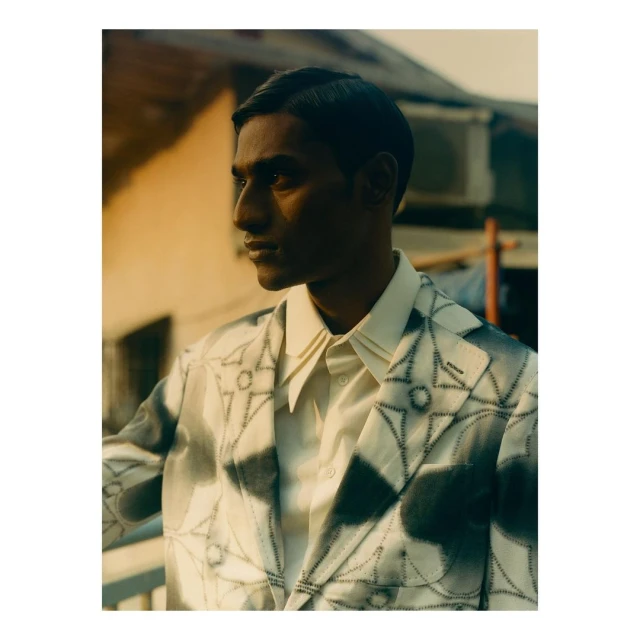 Portrait of a dark-skinned man wearing a patterned blazer and off-white collared shirt, standing outside with dappled lighting.