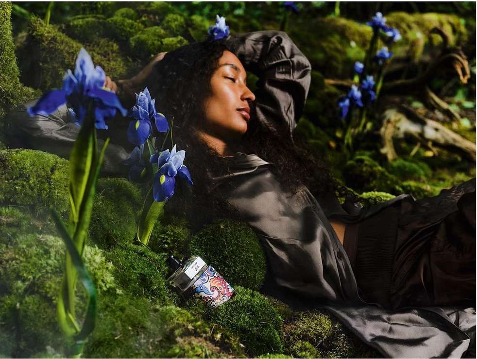 Woman in a flowing grey dress relaxes on moss surrounded by blue iris flowers. Bottle of perfume next to her in an outdoor setting.