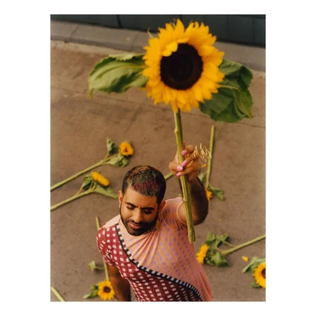 A man wearing a patterned shirt holding a tall sunflower against a neutral concrete ground with scattered sunflowers.