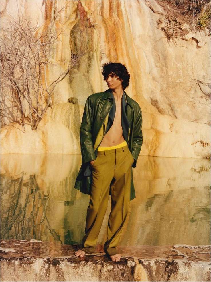 An adult male fashion model in a green jacket and tan trousers strikes a pose outdoors against a textured rock formation and pool.