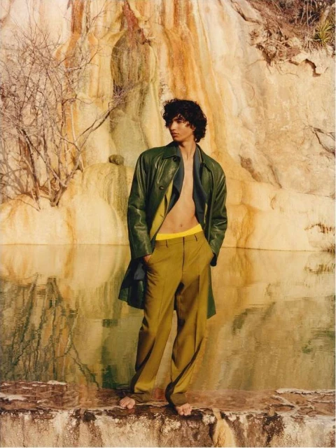 An adult male fashion model in a green jacket and tan trousers strikes a pose outdoors against a textured rock formation and pool.