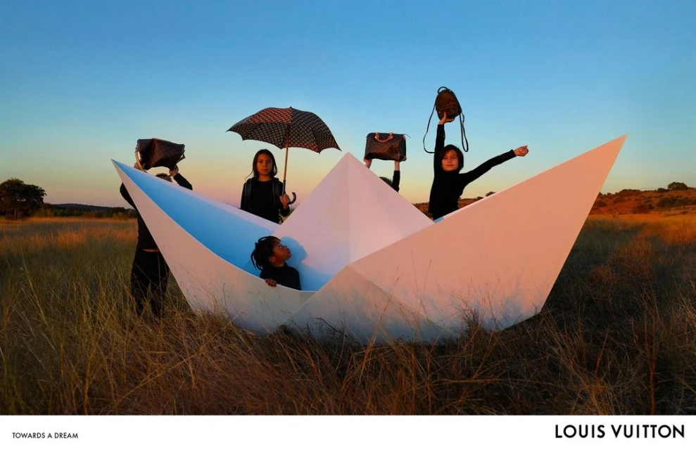 Children inside a large paper boat on a grassy plain at sunset holding Louis Vuitton handbags and an umbrella.  