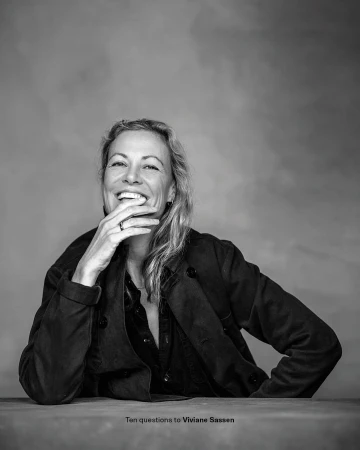 Monochrome portrait of an adult woman in a dark jacket with her hand near her face smiling against a textured studio backdrop