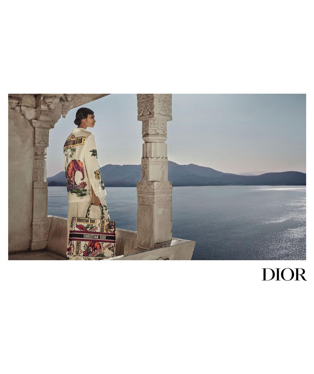 Model wearing printed jacket and pants with Dior tote bag stands overlooking a lake, mountains, and decorative stone columns.