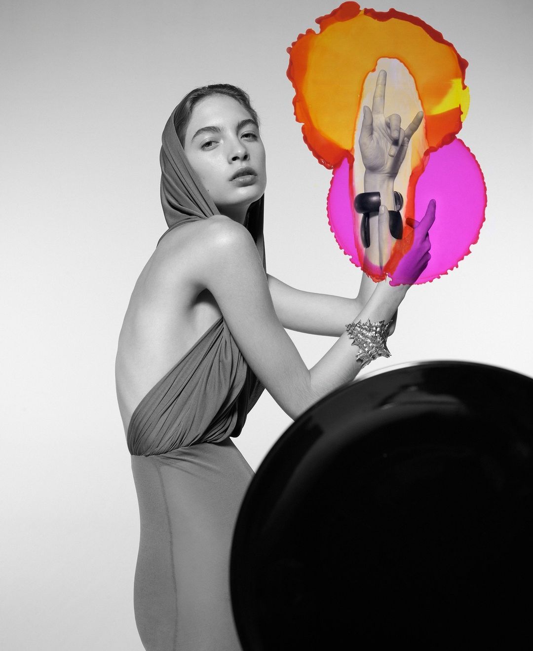 Monochrome portrait of a fashion model in a draped gown with a hood, posing with colorful translucent shapes and hand gestures.