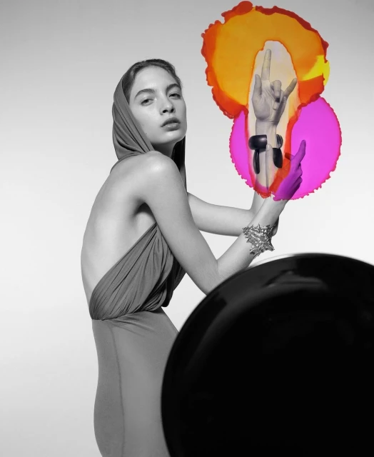 Monochrome portrait of a fashion model in a draped gown with a hood, posing with colorful translucent shapes and hand gestures.