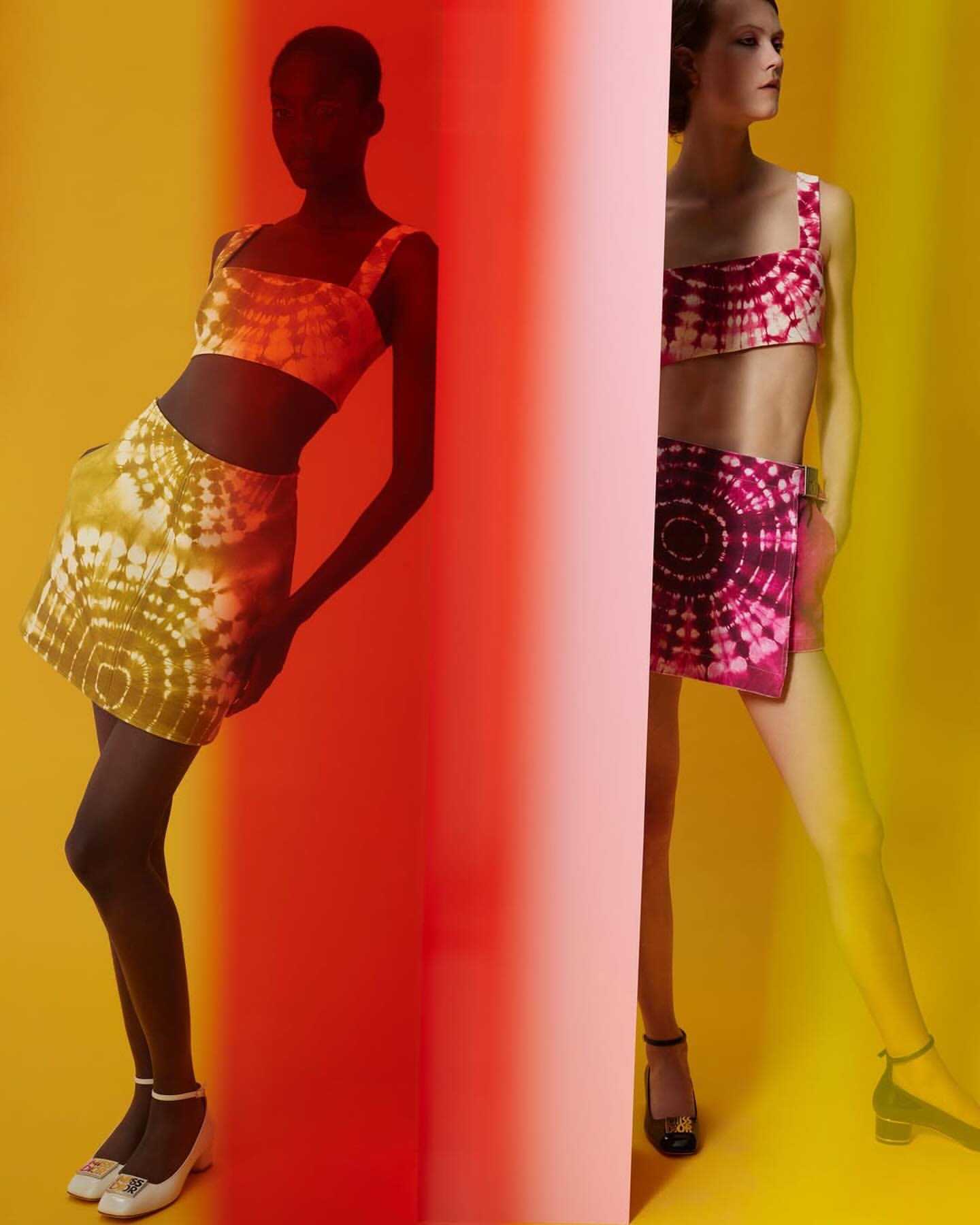 Two women pose in matching tie-dye crop tops and skirts against a vibrant yellow and red backdrop, showcasing a retro fashion aesthetic.