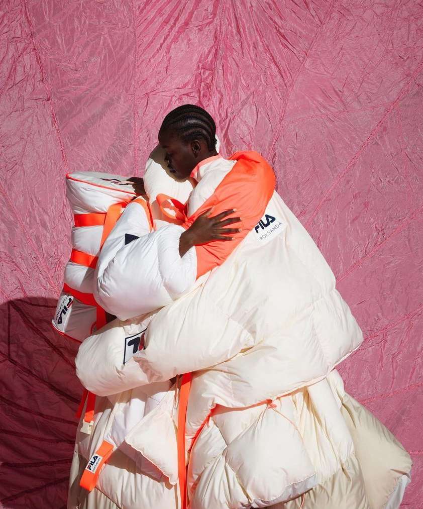 Editorial shot of a Black woman in a voluminous white and orange Fila jacket embracing a pile of puffy Fila-branded garments against a pink backdrop.