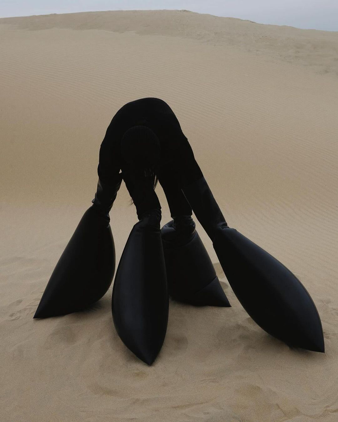 Abstract artistic desert landscape featuring a person in a black suit upside down in the sand dunes. The sky is overcast.
