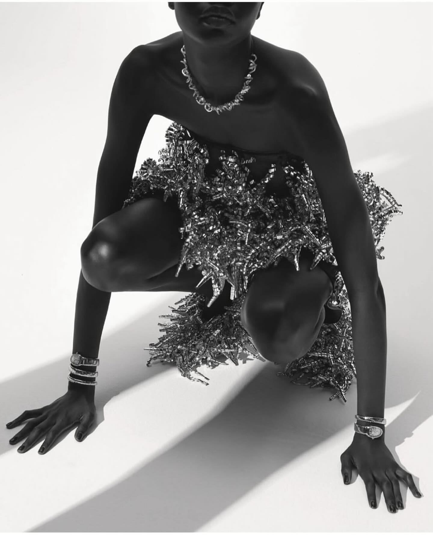 Monochrome portrait of a model in a crouched position wearing jewelry and a metallic dress against a white backdrop.