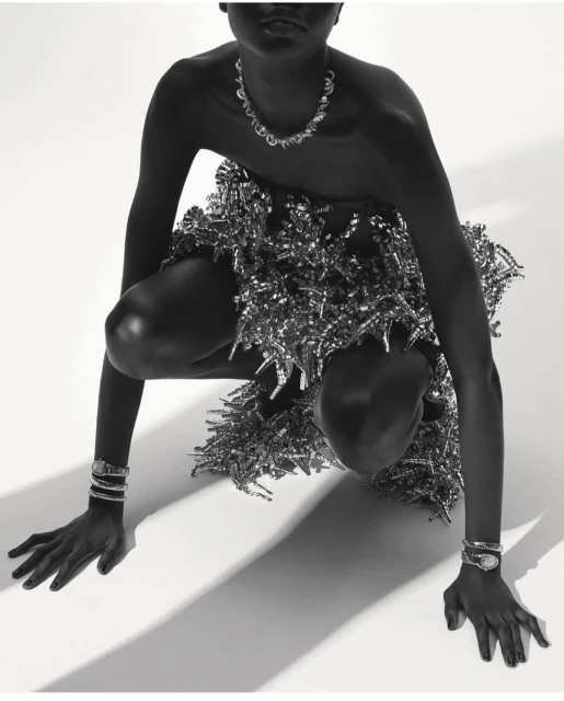 Monochrome portrait of a model in a crouched position wearing jewelry and a metallic dress against a white backdrop.