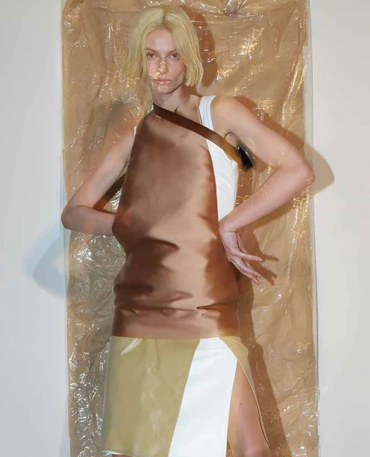 Woman in a sleeveless white and brown dress poses in front of a wrinkled tan backdrop in a fashion or editorial context, studio lighting.