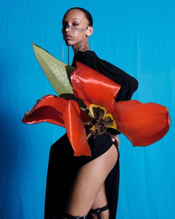 Adult female poses with red flower on a blue backdrop, wearing a black dress with a futuristic wire face accessory.