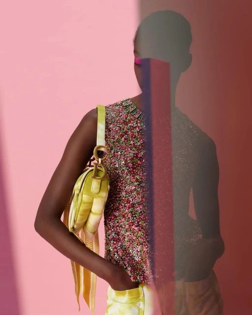 Fashionable woman poses indoors against a pink background with lime green purse and beaded top; an abstract transparent block.