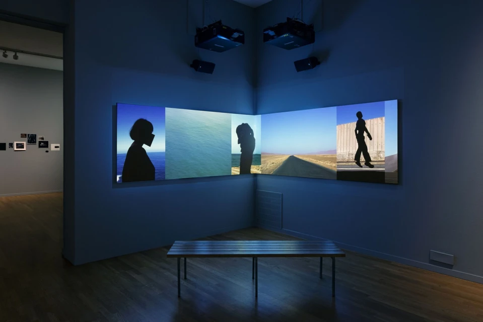 Installation view of a gallery with a multi-panel video display showing silhouette portraits, a coastal scene, and a desert landscape.