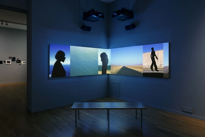 Installation view of a gallery with a multi-panel video display showing silhouette portraits, a coastal scene, and a desert landscape.