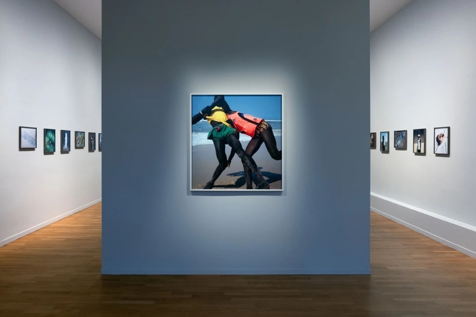 Framed photograph exhibiting two figures in swimsuits wrestling on a beach, presented in a modern art gallery with wooden floors.