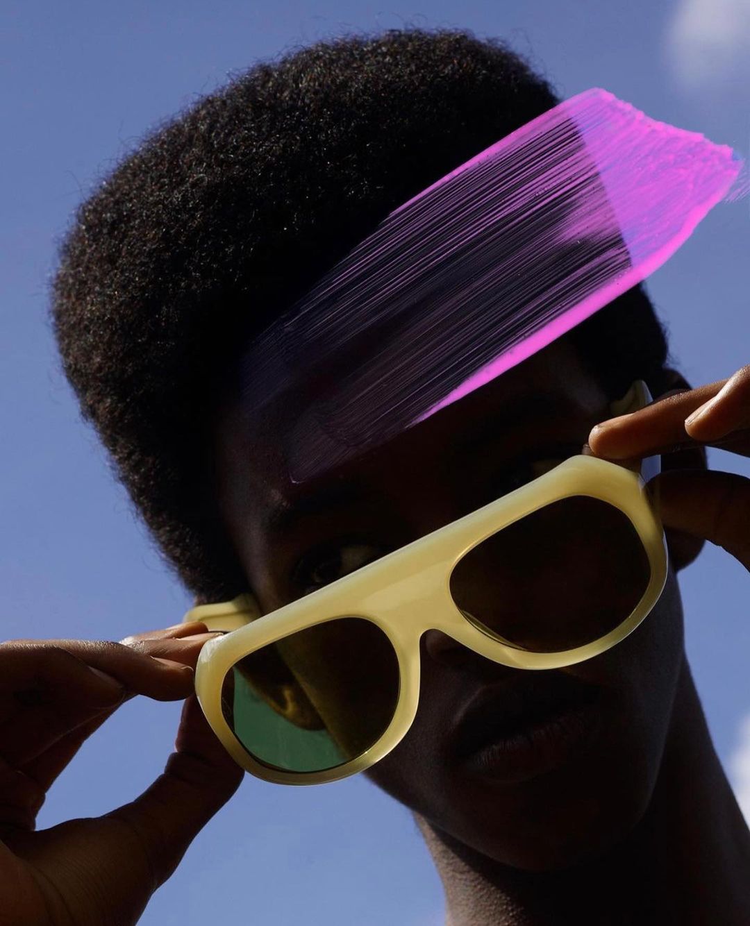 Close-up of a Black person wearing cream-colored sunglasses and looking up at the sky with an overlaid pink brushstroke effect.
