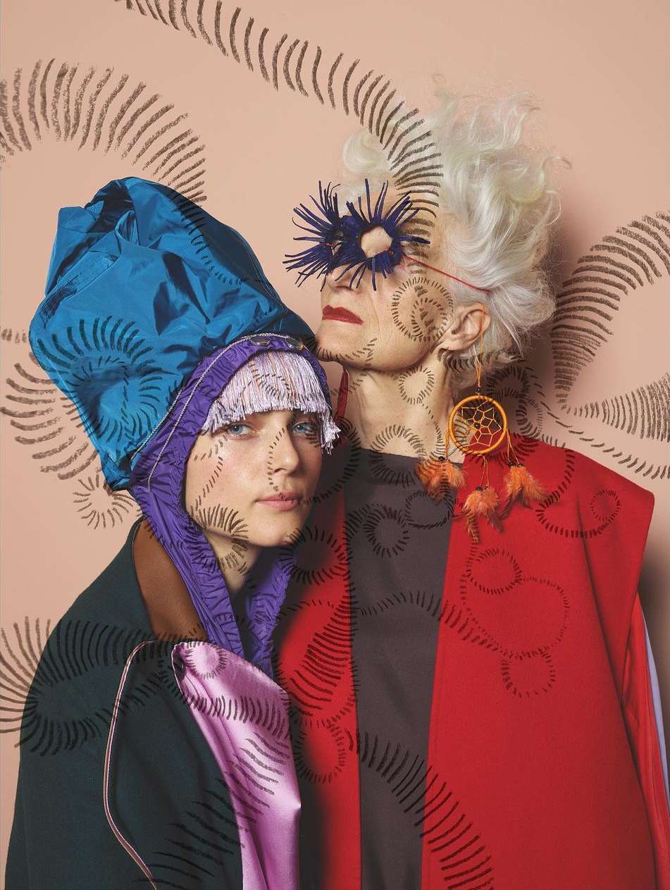 Two women in artistic outfits, one with a blue headpiece and the other with a feathered mask, stand against a peach background with sketched swirls.
