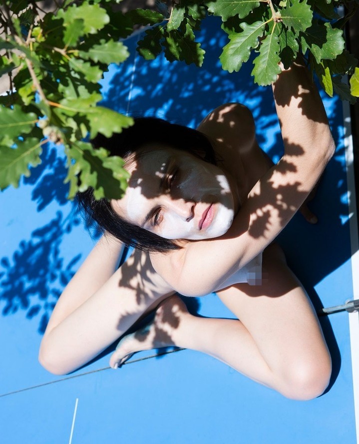 Eye-level portrait of a fair-skinned adult woman lying on a blue surface, face partially obscured by green leaves, bathed in sunlight.