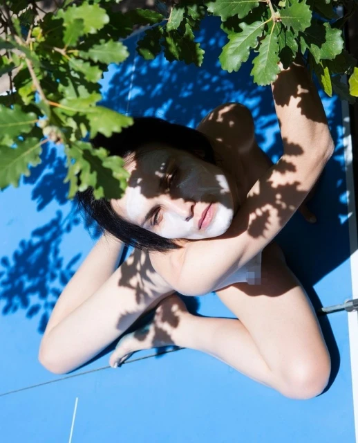 Eye-level portrait of a fair-skinned adult woman lying on a blue surface, face partially obscured by green leaves, bathed in sunlight.