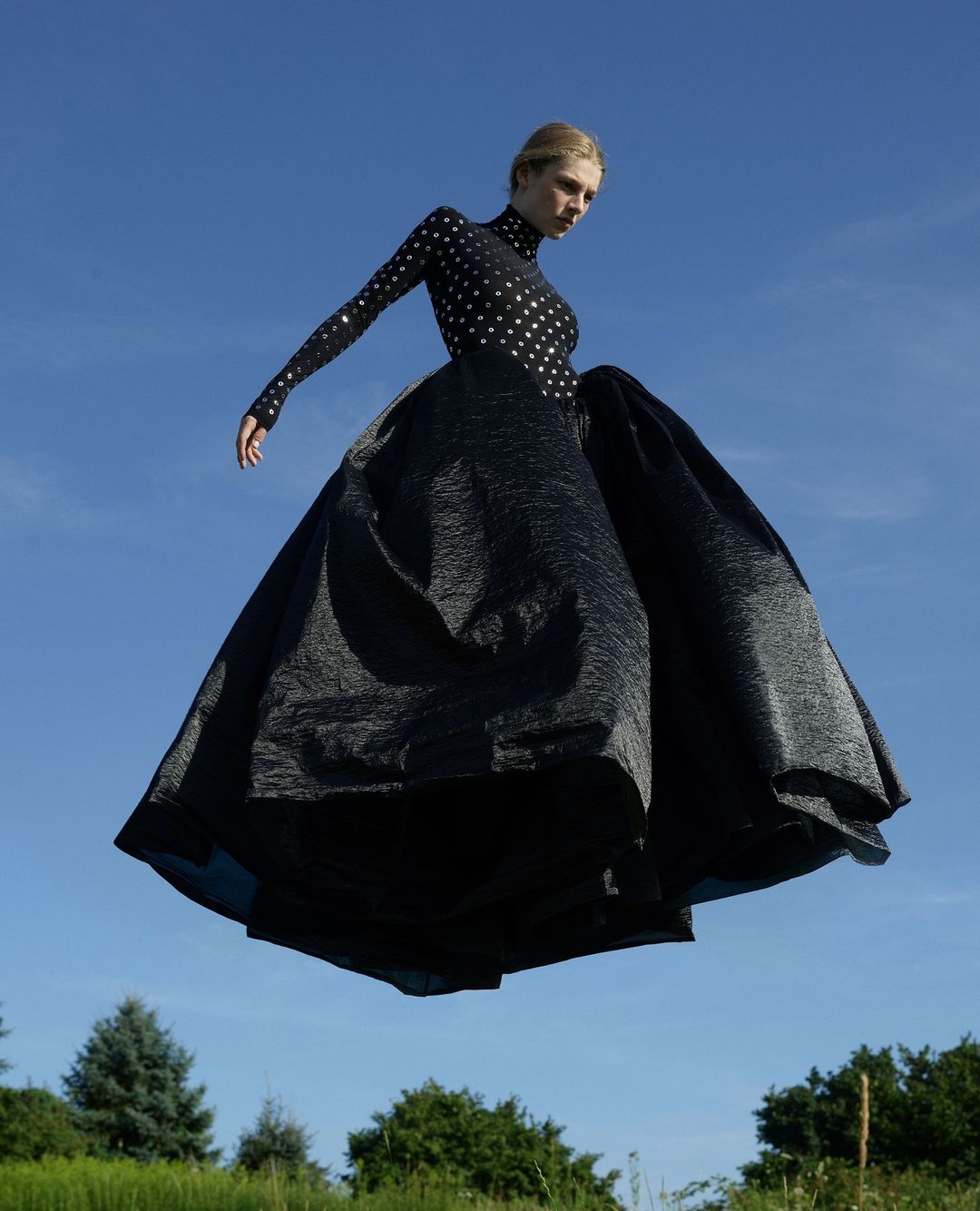 A woman in an elegant black gown floats over a green field against a blue sky, creating a surreal and captivating fashion scene.