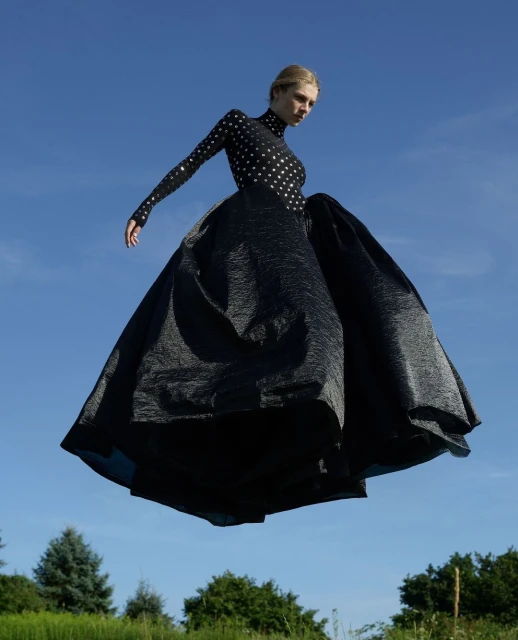 A woman in an elegant black gown floats over a green field against a blue sky, creating a surreal and captivating fashion scene.