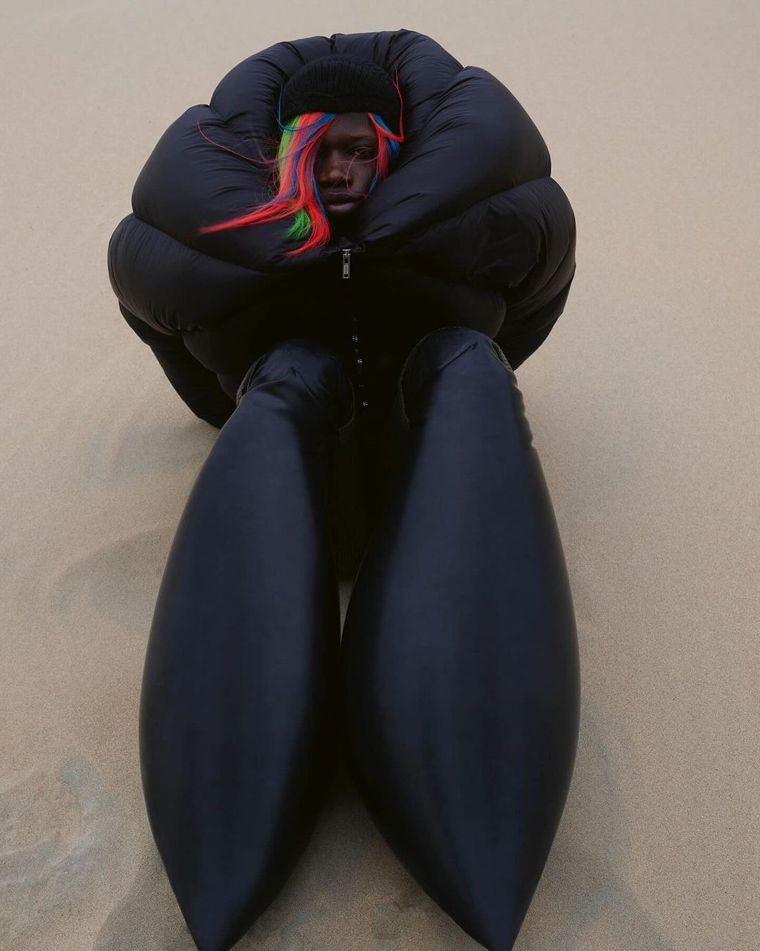 Person in oversized puffer jacket and pants sitting outdoors. The model is wearing a black beanie and has rainbow colored hair.