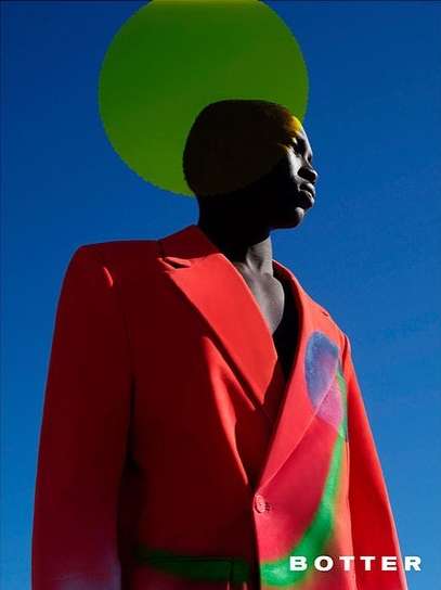 A dark-skinned man in a vibrant red jacket poses in an outdoor fashion shot with a bright blue sky and green graphic circle backdrop.