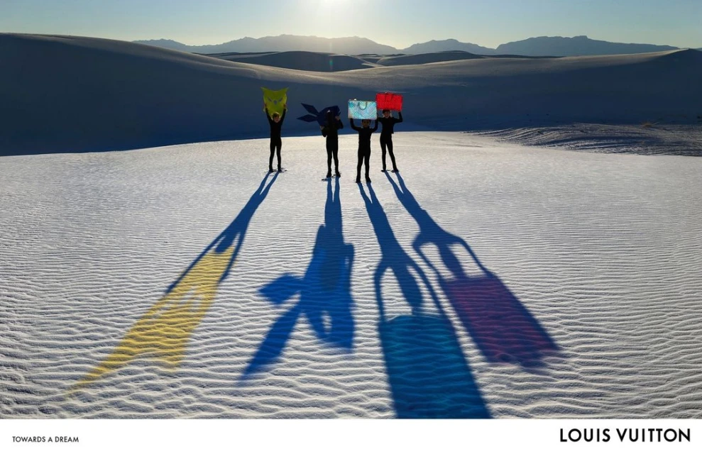 Four people stand in the sand dunes holding up yellow, blue, teal, and red bags with mountains in the background on a sunny day.