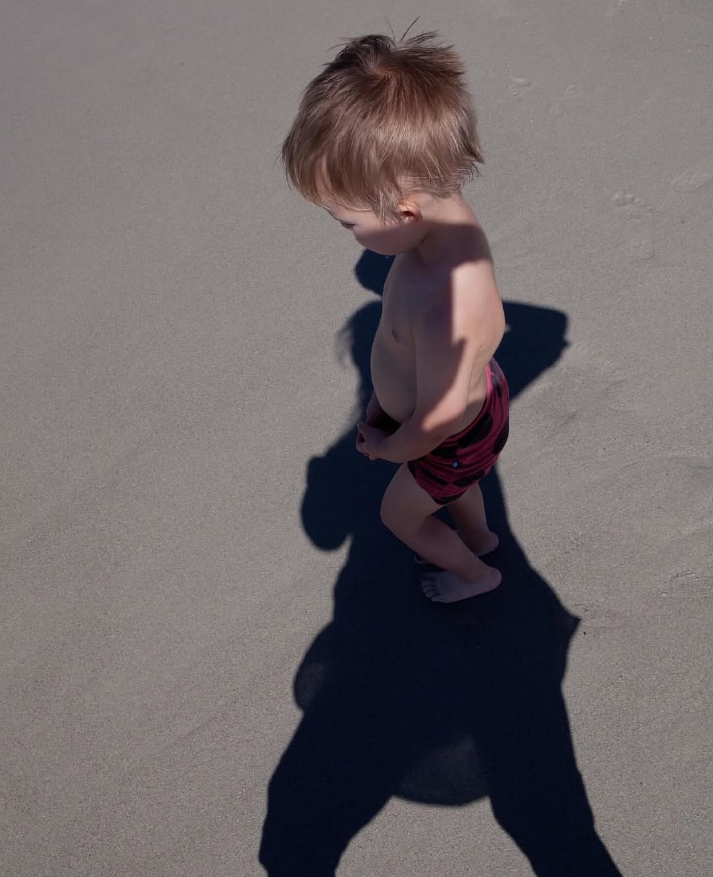 Toddler with blond hair walks along a sandy beach in swimwear, casting a long shadow, enjoying the sun and open space.