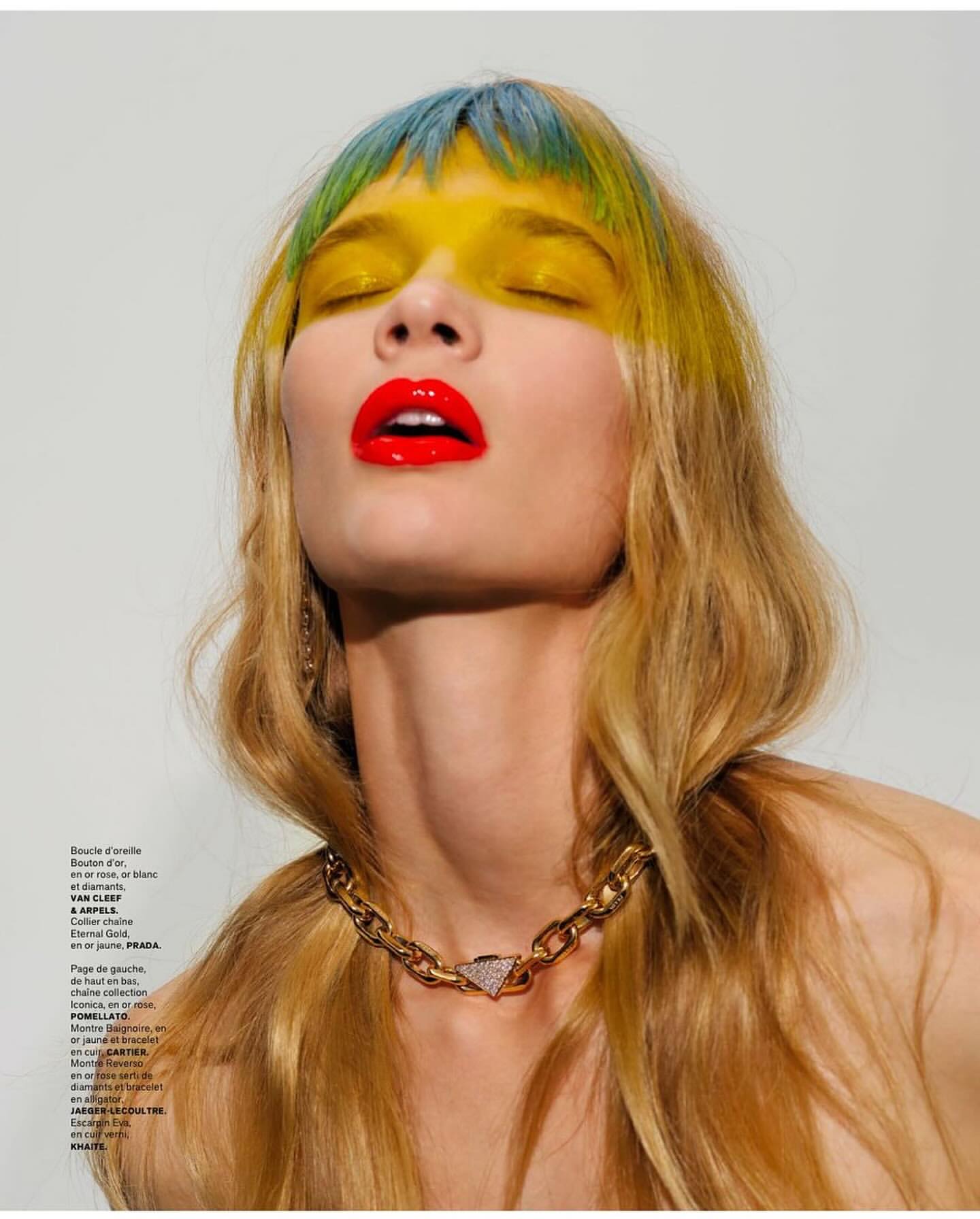 Close-up of a woman with a bold red lip and colorful hair wearing a gold necklace against a light background in a fashion editorial.