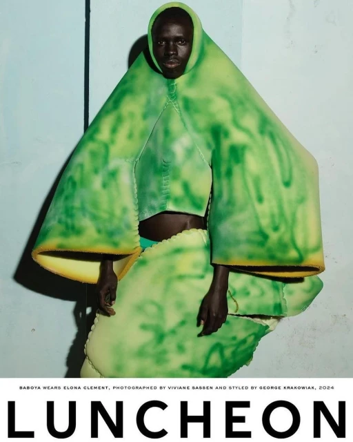 Avant-garde fashion shot featuring a Black model in a green and yellow textured outfit against a pale blue wall. Editorial style.