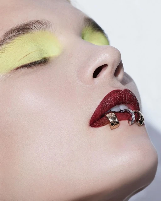 Close-up beauty shot of a woman with creative makeup and lip jewelry against a white backdrop, emphasizing the beauty and fashion elements.